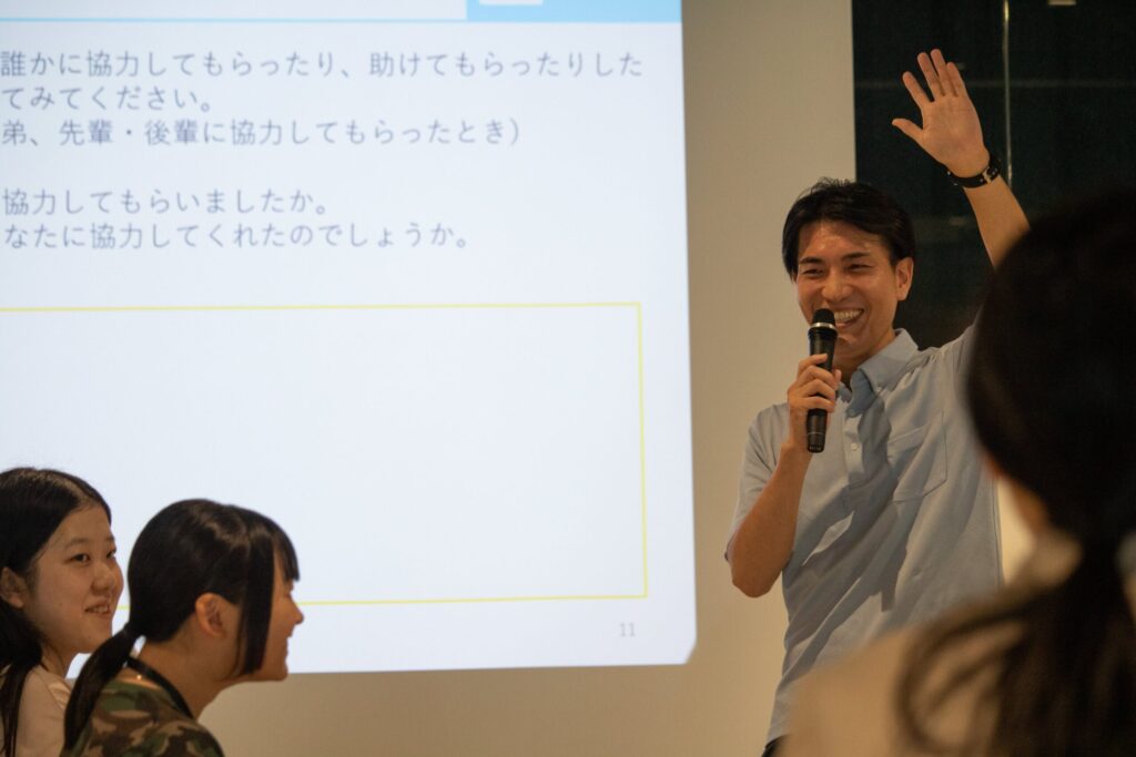 写真。左手を挙げて意見を募る若杉講師。