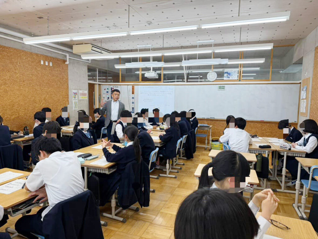 写真。茨城県立水海道第一高等学校附属中学校の教室で、生徒の中を歩く代表濱。