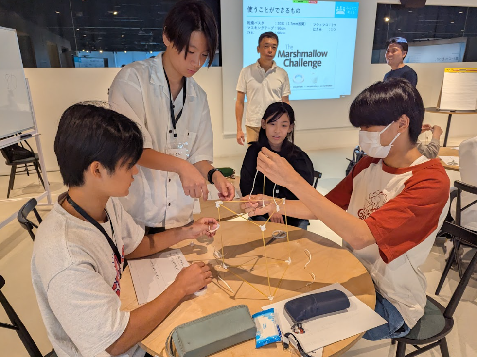 写真。スパゲッティーとマシュマロを使って中高生4名がワークショップをしている。それを講師2名が見守っている。