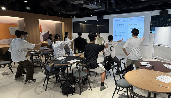 写真。受講生らが立ち上がり、手を前に出してポーズを取っている。